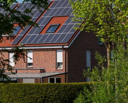 A modern brick house with solar panels on the roof, surrounded by lush greenery, under a clear blue sky.